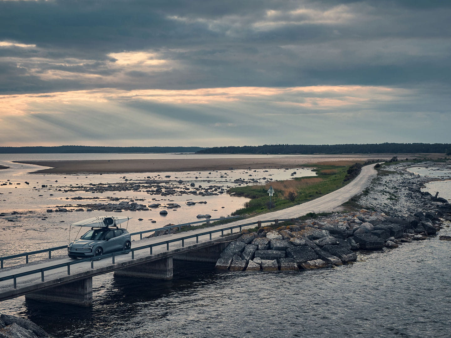 Grå helt elektrisk EX30 med en kajak på taket kör över en bro i skymningsljus.