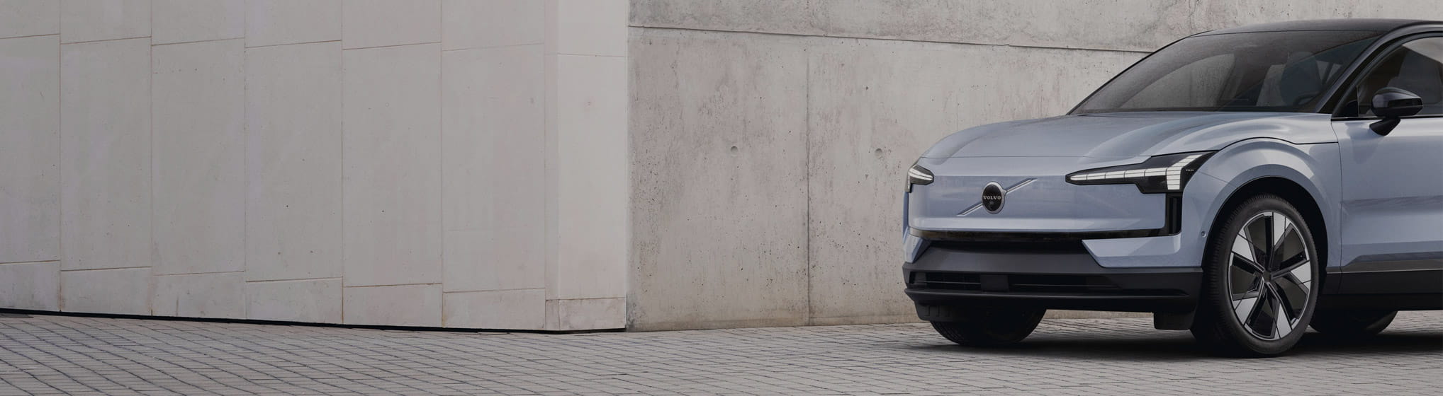 Ljusblå helt elektrisk EX30 parkerad på kullerstensgata framför en grå betongvägg.